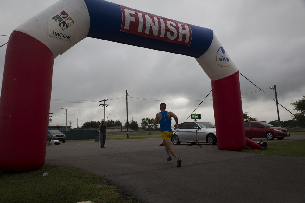 A runner in a blue tank top and yellow shorts is approaching the finish line of a race under an inflatable red, white, and blue 'FINISH' arch. The arch displays logos for IMCOM and MWR, suggesting this is a military community event. A race official or photographer stands in the background, and a timing clock is visible near the finish line.