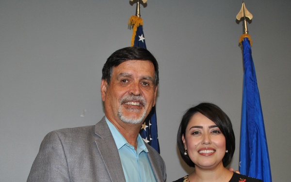 Two individuals pose together in what appears to be an official military or government ceremony setting. The woman on the right is holding a coin/medallion and wearing a medal on her chest, along with a blue folder, suggesting she is the recipient of an award. Two flags — an American flag and what appears to be a U.S. Air Force flag — are visible in the background.