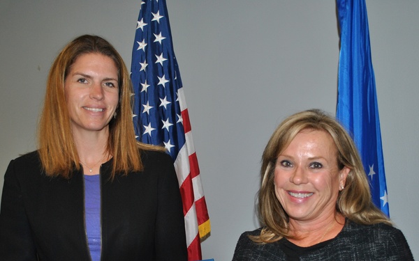 Two women are posing together at what appears to be a formal recognition ceremony. The woman on the left is holding a Certificate of Service awarded to 'Leslie C. Brown' in recognition of Twenty Years of service in the Government of the United States of America. An American flag and a light blue flag (likely U.S. Air Force) are visible in the background.