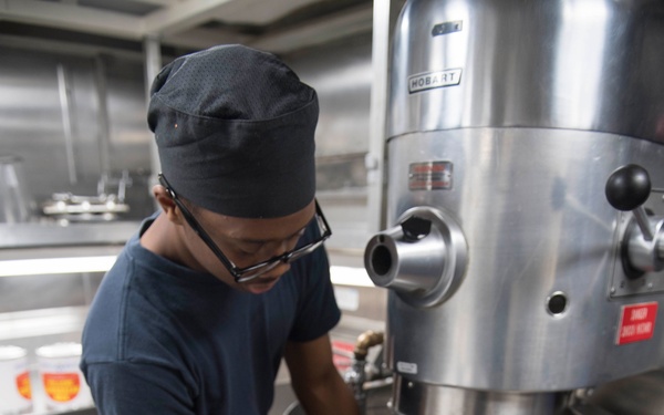 A kitchen worker wearing a dark blue uniform and skull cap is pouring an ingredient from a container into a large commercial Hobart stand mixer bowl. The setting appears to be a professional or institutional kitchen with stainless steel equipment and shelving. The worker is wearing glasses and appears focused on the task of adding what looks like a dry ingredient, possibly flour or sugar.