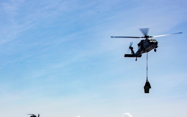 Two military helicopters are conducting a vertical replenishment (VERTREP) operation over open ocean near a large white hospital ship identifiable by red cross markings on its hull. The foreground helicopter is carrying a slung load suspended beneath it via a cargo hook. The scene depicts a coordinated naval logistics operation in clear daylight conditions.
