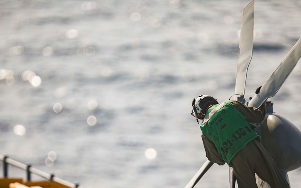 The image shows a close-up view of a helicopter rotor hub and blades on what appears to be a naval vessel's flight deck, with the open ocean visible in the background. A green tag or label is attached to the rotor hub assembly, and a flight suit or coverall garment is draped over the rotor mast. The ship's deck edge, safety railings, and tie-down equipment are visible in the left portion of the frame.