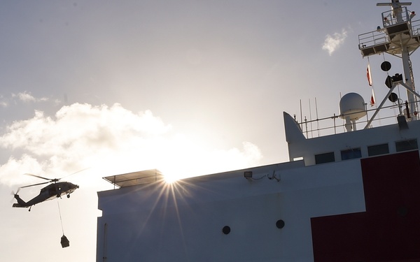 A military helicopter, likely a Sikorsky MH-60 Seahawk, is performing a vertical replenishment or cargo transfer operation near a large white hospital ship marked with a prominent red cross. The scene is captured at low sun angle, creating a dramatic starburst lens flare effect. The ship appears to be underway at sea, consistent with a U.S. Navy hospital ship such as the USNS Comfort or USNS Mercy.