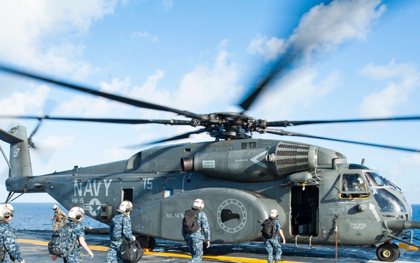 A U.S. Navy helicopter, marked 'NAVY' and 'HM-15' with the number 75, is parked on a ship's flight deck with its rotors spinning. Several Navy personnel in blue digital camouflage uniforms and white flight deck helmets are walking toward the open helicopter door. The scene takes place at sea under partly cloudy skies.