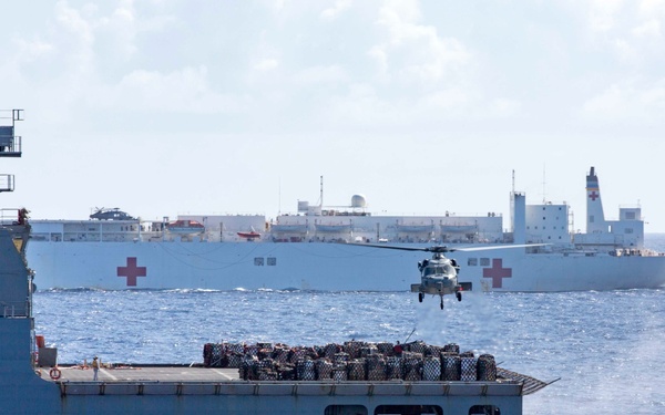 A naval logistics operation is underway at sea, with a military cargo/supply ship in the foreground loaded with palletized cargo. A helicopter is conducting a vertical replenishment (VERTREP) operation between the supply vessel and a large white hospital ship visible in the background, identifiable by prominent red cross markings. The scene depicts a coordinated underway replenishment (UNREP) mission in open ocean conditions.
