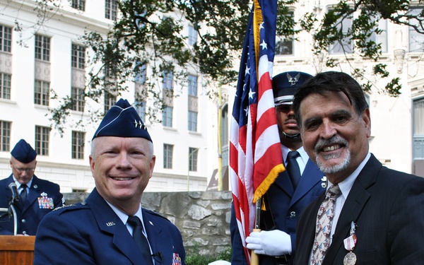 A U.S. Air Force officer identified by nameplate as 'Thompson' presents an 'Outstanding Civilian Career Service Award' from the Department of the Air Force to a civilian named 'Randy E. Brown' at a ceremony held at The Alamo in San Antonio, Texas. Both men are smiling and holding the certificate together. An honor guard member holds the American flag in the background, and another uniformed officer speaks at a podium.
