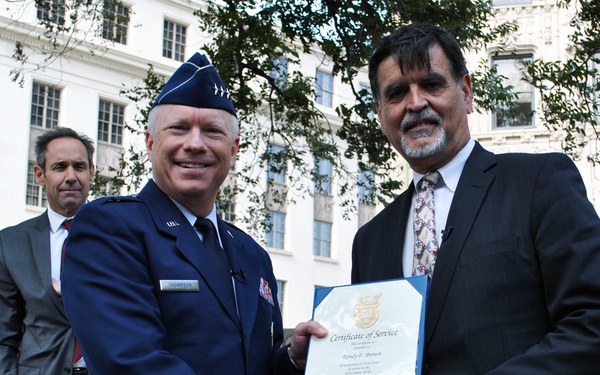 A U.S. Air Force officer is presenting a Certificate of Service to a civilian man during an outdoor ceremony. The two men are shaking hands while jointly holding the framed certificate. A third man in a suit stands in the background near a podium.