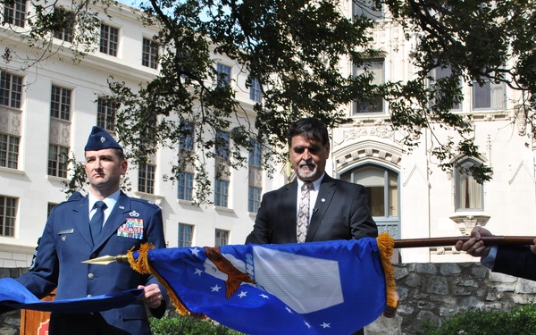 An outdoor military ceremony is taking place in front of a large white neoclassical building. Two uniformed U.S. Air Force personnel and one civilian in a dark suit are handling what appears to be a U.S. Air Force flag with gold fringe. The setting includes a stone courtyard with a wooden podium, suggesting a formal presentation or retirement ceremony.