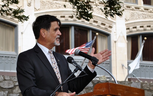 A man in a dark suit and patterned tie is speaking at a wooden podium bearing 'The Alamo - The Cradle of Texas Liberty' logo. The event appears to be an official or ceremonial occasion held outdoors at or near the Alamo in San Antonio, Texas. An American flag and a white flag are visible in the background, along with a historic multi-story building.