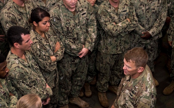 A military gathering showing a senior Army officer in OCP (Operational Camouflage Pattern) uniform addressing a large group of Navy personnel in NWU (Navy Working Uniform) digital camouflage. The scene appears to be an informal briefing or all-hands meeting, with the Army officer at the center of a circle of seated and standing sailors. The setting appears to be an indoor military facility or hangar.