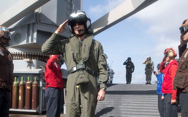 A military pilot in an olive green flight suit and helmet is saluting on the deck of what appears to be an aircraft carrier or naval vessel. Crew members in various colored vests and gear are lined up on both sides, also saluting, suggesting a formal honors ceremony or distinguished visitor protocol. The scene takes place on an outdoor flight or weather deck with naval equipment visible in the background.