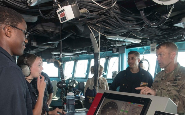 The image shows a group of military personnel on what appears to be the bridge of a naval vessel. A sailor in dark navy coveralls is standing at a navigation/control console while a person in Army combat uniform appears to be receiving a briefing or tour. Several other uniformed personnel are visible in the background near the bridge windows.
