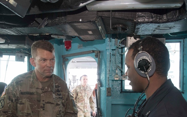A U.S. Army soldier with the name tag 'Buchanan' and rank insignia visible is engaged in conversation with a naval crew member wearing headphones inside what appears to be a ship's bridge or control room. A second soldier in camouflage uniform is visible in the background near a doorway. The setting appears to be a military vessel with navigation and communication equipment visible.