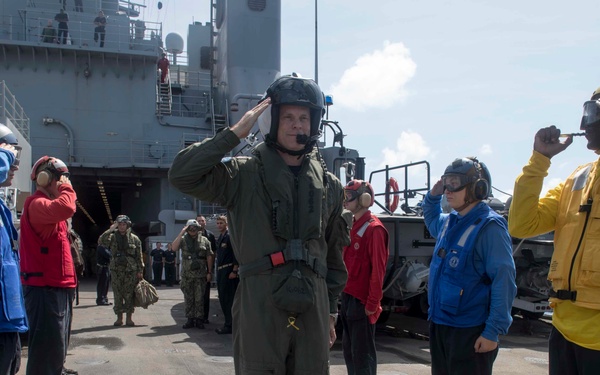 A military ceremony is taking place on the flight deck of a naval vessel, with personnel in various colored safety vests and flight gear rendering salutes. A central figure in an olive green flight suit and helmet with a communications microphone is saluting, flanked by crew members in blue, red, and yellow jerseys also saluting. The scene appears to be a formal honors or arrival/departure ceremony at sea, with a larger ship visible in the background.