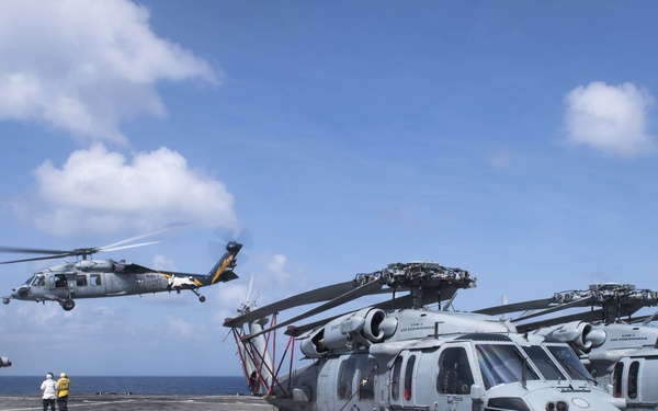 The image shows a naval vessel's flight deck with multiple military helicopters parked and one in flight. Several grey helicopters, appearing to be MH-60 Seahawk variants, are secured on the deck while one is airborne nearby. Deck crew members in colored safety vests are visible on the flight deck overseeing operations.