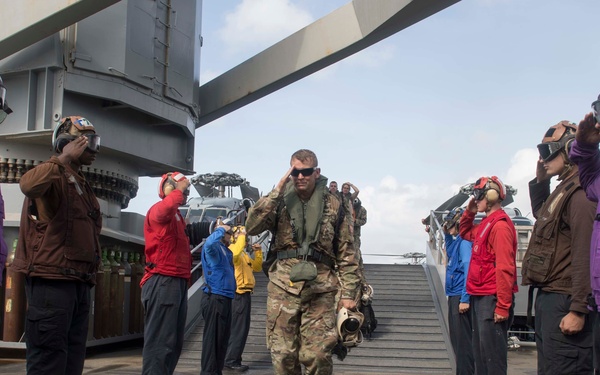 A military personnel in camouflage uniform and flight gear walks across a naval vessel's deck while being rendered salutes by a formation of sailors in colored jerseys. The scene appears to be a formal honors ceremony, likely welcoming or departing a distinguished visitor or high-ranking officer. The setting is clearly a warship flight deck, with naval superstructure and equipment visible in the background.