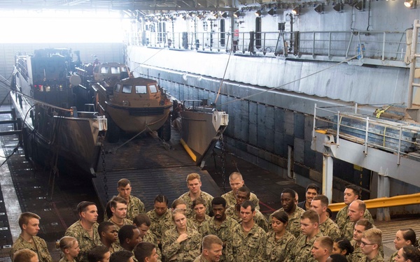 A large group of U.S. Navy personnel in digital camouflage (NWU Type I) uniforms are assembled in the well deck of an amphibious warfare ship. The gathering appears to be a briefing or all-hands meeting, with personnel standing in a semi-circular formation facing a central speaker. Landing craft, likely LCUs (Landing Craft Utility), are visible secured in the flooded well deck behind the assembled sailors.