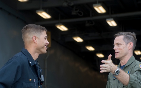 Two military personnel are engaged in conversation aboard what appears to be a naval vessel, likely an aircraft carrier hangar deck. One individual is wearing dark navy coveralls and the other is wearing an olive green flight suit with a U.S. flag patch on the sleeve. The setting features industrial overhead lighting and metal structures typical of a ship's interior bay.