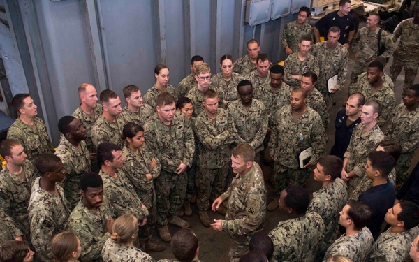 A large group of military personnel, predominantly wearing U.S. Navy NWU (Naval Working Uniform) camouflage pattern uniforms, are gathered in a circle formation in what appears to be a ship's hangar bay or large interior compartment. A central figure, appearing to wear Army-style camouflage, is addressing the assembled group. The gathering resembles a briefing, all-hands meeting, or command address.