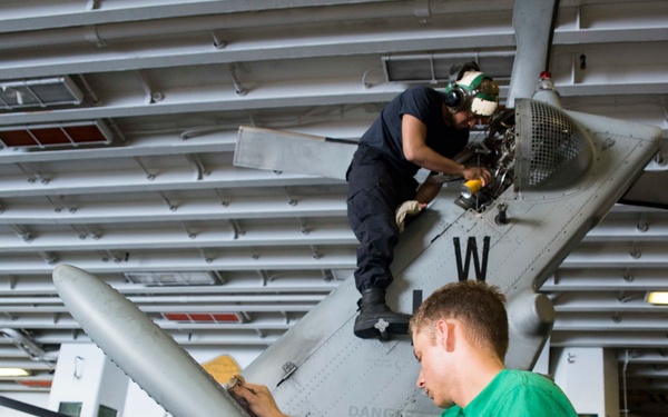 Two maintenance technicians are working on a military aircraft inside what appears to be a naval vessel's hangar bay. One technician in a green shirt works on the lower wing/fuselage area while another in dark clothing and hearing protection works on the upper section near the engine or rotor hub. The setting is clearly an enclosed shipboard hangar with overhead lighting and safety signage visible.