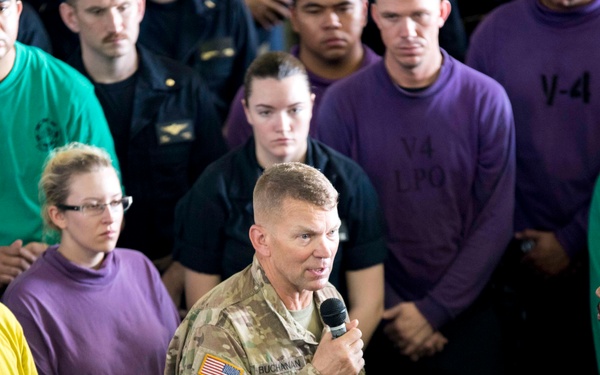 A U.S. Army officer in camouflage uniform is addressing a large group of military personnel and civilians while holding a microphone. The speaker, whose name tape reads 'BUCHANAN,' wears a 10th Mountain Division patch and is gesturing with an outstretched hand. The audience surrounds him in a semicircle, wearing various colored shirts indicating different unit or division assignments.