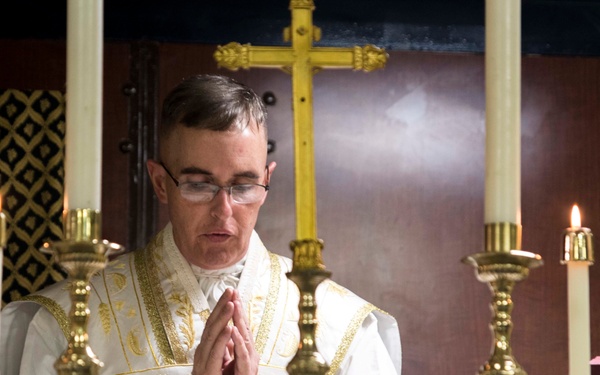 A Catholic priest in white and gold vestments stands at an altar with hands clasped in prayer. He is surrounded by multiple brass candlesticks with lit candles, and a gold crucifix is visible in the background. The setting appears to be a traditional Catholic Mass or liturgical ceremony.