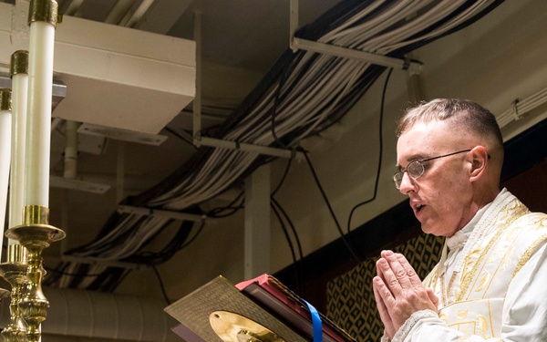 A Catholic priest in white and gold liturgical vestments stands at an altar in prayer with hands clasped together. He is reading from a missal or liturgical book mounted on a brass stand, surrounded by tall white candles in ornate brass candlesticks. The setting appears to be a makeshift or temporary chapel, possibly aboard a military vessel or in an industrial space, given the exposed pipes and cable trays visible in the ceiling.