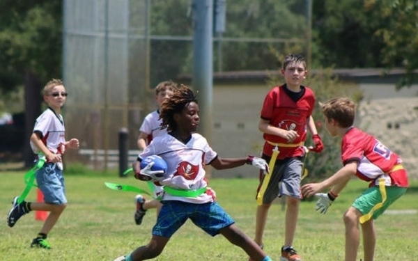 A group of young children are playing flag football on a grass field during what appears to be a sunny day. The central player in a white jersey is carrying a football and attempting to evade defenders who are trying to pull his flags. The scene appears to be an organized youth league or camp setting.