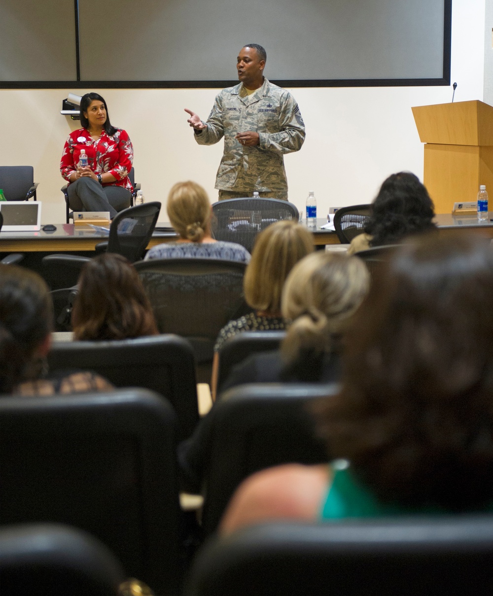 A U.S. Air Force senior non-commissioned officer in ABU (Airman Battle Uniform) camouflage is addressing an audience in what appears to be a conference or briefing room. A woman in a red floral blouse is seated behind him, likely a co-presenter or guest. The audience consists primarily of women seated in rows of black chairs facing the front.