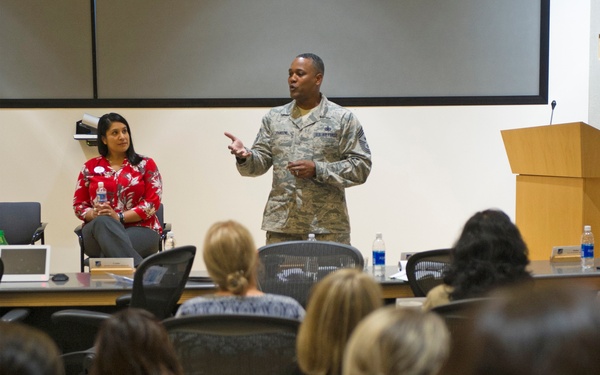 A U.S. Air Force senior non-commissioned officer in ABU (Airman Battle Uniform) camouflage is addressing an audience in what appears to be a conference or briefing room. A woman in a red floral blouse is seated behind him, likely a co-presenter or guest. The audience consists primarily of women seated in rows of black chairs facing the front.