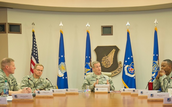 A formal military briefing or conference is taking place in what appears to be a U.S. Air Force command conference room. Several uniformed Air Force personnel are seated around a large wooden conference table, with name placards visible in front of each participant. The room features multiple Air Force unit flags, a unit crest on the wall, and a framed portrait, indicating a high-level command setting.