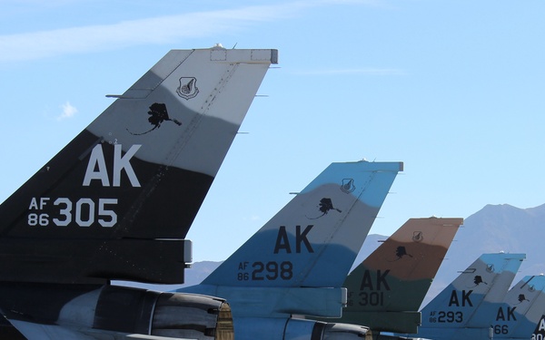 A row of U.S. Air Force F-16 Fighting Falcon jets are parked on a flight line, all bearing 'AK' tail codes indicating Alaska Air National Guard assignment. Each aircraft features a distinctly colored tail fin with unique camouflage or scenic paint schemes. Ground support equipment is visible beneath the aircraft, suggesting active maintenance or pre-flight operations.