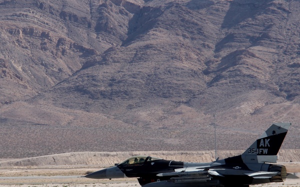 A U.S. Air Force F-16 Fighting Falcon jet fighter is taxiing on a runway in a desert environment. The aircraft bears tail markings 'AK' and '354 FW', identifying it as belonging to the 354th Fighter Wing based in Alaska. Arid mountain terrain is visible in the background, consistent with a Nevada desert installation such as Nellis Air Force Base.
