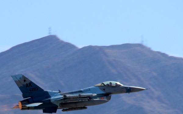 A U.S. Air Force F-16 Fighting Falcon jet fighter is captured in flight against a backdrop of arid, brown mountain terrain under a clear blue sky. The aircraft bears 'AK' tail codes indicating it belongs to an Alaska Air National Guard unit, with a partially visible serial number appearing to read '293'. The jet is in a climbing or level flight attitude with afterburner engaged, visible as an orange glow at the engine exhaust.