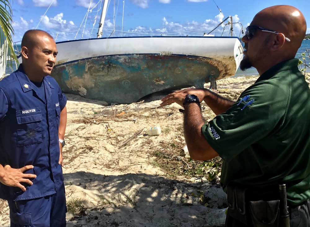 Two officials are standing on a beach in front of a damaged, grounded sailboat that appears to have been washed ashore, likely after a storm or hurricane. One individual is wearing a blue U.S. Coast Guard uniform with the name tag 'NGUYEN,' while the other is wearing a green uniform with 'DPNR' insignia (Department of Planning and Natural Resources). The man in green is gesturing toward the vessel, apparently discussing its condition or the assessment process.