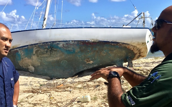 Two officials are standing on a beach in front of a damaged, grounded sailboat that appears to have been washed ashore, likely after a storm or hurricane. One individual is wearing a blue U.S. Coast Guard uniform with the name tag 'NGUYEN,' while the other is wearing a green uniform with 'DPNR' insignia (Department of Planning and Natural Resources). The man in green is gesturing toward the vessel, apparently discussing its condition or the assessment process.