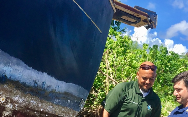 Two officials are inspecting a grounded or beached vessel, with one holding documents and appearing to take notes. The boat shows significant hull damage, corrosion, and marine growth, suggesting it has been out of water or abandoned for some time. The setting appears to be a coastal or shoreline area with dense vegetation in the background.
