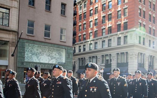 A formation of U.S. Army soldiers in dress blue uniforms marches in a military parade along a city street. The procession appears to be taking place in a major urban area, likely New York City, based on the architecture and street layout visible in the background. Soldiers are marching in organized columns under bright daylight conditions.