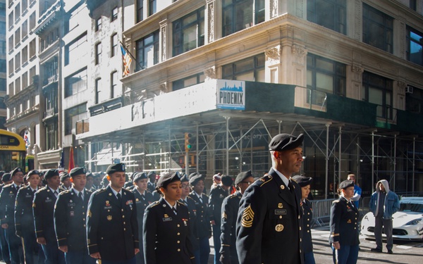 A formation of U.S. Army soldiers in dress uniforms marches in a parade along a city street, likely in New York City based on the urban architecture and a yellow bus marked 'FERRY' visible in the background. The soldiers are wearing Army Service Uniforms (ASUs) with black berets, and a senior non-commissioned officer (identifiable by chevron rank insignia consistent with a Sergeant Major) leads the formation. The scene appears to be a formal military parade, possibly a Veterans Day or Memorial Day event.