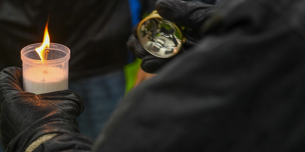 A close-up photograph showing two individuals wearing black gloves at what appears to be a candlelight vigil or memorial gathering. One person holds a lit candle in a small plastic/wax holder, while the other holds a reflective metallic object, possibly a bell or lid. The dark clothing and candlelight suggest a solemn or ceremonial occasion.