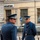NATO Military Committee visit to Sarajevo Two military or ceremonial guard personnel in matching blue dress uniforms are standing outdoors in what appears to be a European city setting. The foreground figure faces away from the camera while the background figure faces forward, both wearing peaked caps with insignia and white gloves. A bank building with a partially visible 'UniCredit Bank' sign is visible in the background.