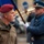 NATO Military Committee visit to Sarajevo A military officer wearing a maroon beret and a name tag reading 'Petr PAVEL' with four stars on his collar stands in the foreground at what appears to be a formal military ceremony. In the background, a uniformed soldier in a blue dress uniform and peaked cap holds a flag pole while a brass instrument player is also visible. The setting appears to be an outdoor ceremonial event, likely in a European city.
