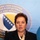 NATO Military Committee visit to Sarajevo A woman is speaking at a podium with microphones in front of an official backdrop displaying the coat of arms and text referencing Bosnia and Herzegovina. She is wearing a black blazer with a patterned scarf and small earrings. The setting appears to be a formal press conference or official government event.