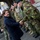 NATO Military Committee visit to Sarajevo A civilian woman in a navy blue coat is shaking hands with a soldier wearing Dutch military camouflage uniform, identifiable by the Netherlands flag patch on the sleeve. Several military personnel in various camouflage uniforms are lined up in the background, suggesting an official greeting or inspection ceremony. The setting appears to be a European courtyard or building entrance with an arched passageway visible.