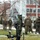 NATO Military Committee visit to Sarajevo A tracked ground robot, consistent with an EOD (Explosive Ordnance Disposal) unmanned ground vehicle, is positioned in the foreground on a grassy area. In the background, several military or security personnel are visible, including one individual wearing a bomb disposal suit. The setting appears to be an outdoor training exercise or demonstration in a cold-weather environment.