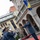 NATO Military Committee visit to Sarajevo A military band ceremony is taking place outdoors in front of a historic European-style building. A conductor in a blue military uniform directs musicians, including a clarinetist in the foreground, while uniformed soldiers stand in formation. The flag of Bosnia and Herzegovina is prominently displayed on a flagpole, suggesting this is an official state or military ceremony.