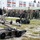 NATO Military Committee visit to Sarajevo The image shows a military demonstration featuring two EOD (Explosive Ordnance Disposal) robots positioned in the foreground on a grassy, partially snow-dusted field. A large group of military personnel in various camouflage uniforms are gathered in the background observing the demonstration. The setting appears to be a military installation or base, with a multi-story building visible in the background.