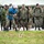 NATO Military Committee visit to Sarajevo A military working dog demonstration is taking place outdoors in wet/rainy conditions. A handler in MARPAT camouflage controls a Belgian Malinois while a person wearing a blue bite suit and protective face shield acts as the decoy/agitator. A multinational group of military observers in various uniforms watches the demonstration from behind a yellow boundary line.