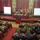 NATO Military Committee visit to Sarajevo A formal military conference is taking place in an ornate hall, identified by the projected slides as a 'Visit of the NATO Military Committee,' dated 24 November 2017. A panel of four military officials sits at a head table on a raised stage, flanked by numerous national flags of NATO member states. The audience consists of dozens of military personnel in various uniforms seated at burgundy-draped tables.