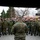 NATO Military Committee visit to Sarajevo A large group of military personnel from multiple nations are gathered outdoors in what appears to be a military installation or compound. A soldier in US Marine Corps digital camouflage (MARPAT) is seen from behind, apparently addressing or briefing the assembled multinational group. The setting appears to be a cold-weather environment based on bare trees and overcast skies, consistent with a Balkans or Eastern European location.